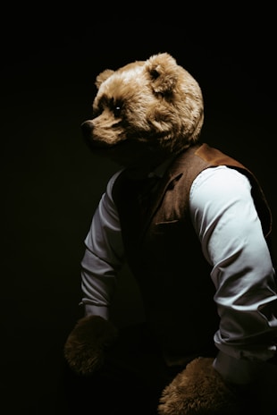 A person wearing a bear costume, complete with a bear head and paws, is dressed in formal attire including a white shirt and a brown waistcoat. The lighting is dramatic, casting shadows and creating a mysterious and theatrical atmosphere.