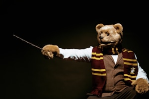 A person is dressed in a bear costume, wearing a maroon and gold scarf, and holding a wooden wand. The costume includes a formal outfit with a white shirt and vest, creating an intriguing combination of whimsical and formal attire.