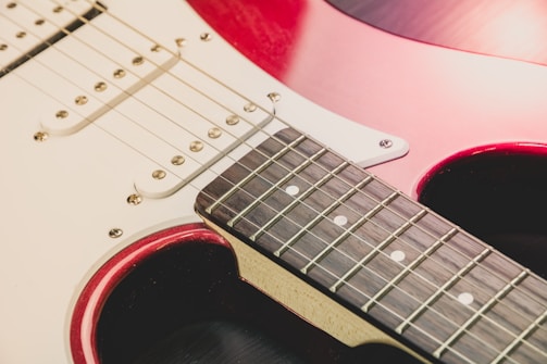 A close-up view of an electric guitar featuring the neck and part of the body. The guitar has metallic strings, a glossy red finish, and a white pickguard. The fingerboard is dark wood with white fret markers.