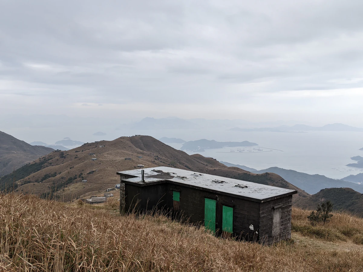 A small hut with green doors on a grassy ridge on Lantau Island, with hills and islands in the haze behind