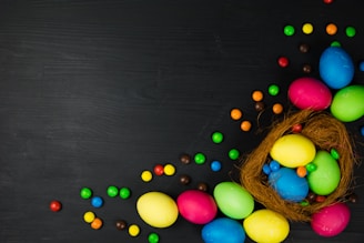A collection of colorful Easter eggs arranged in a nest made of natural fibers, with scattered multicolored candy pieces on a dark surface.