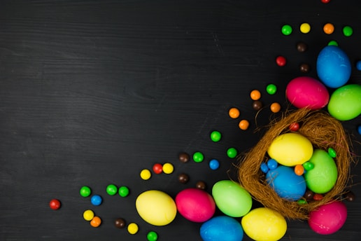 A collection of colorful Easter eggs arranged in a nest made of natural fibers, with scattered multicolored candy pieces on a dark surface.