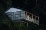 A small, rustic cabin is perched on a hillside surrounded by dense forest. The cabin features a corrugated metal roof and a side wall made of translucent panels. A row of windows, framed by decorative columns, faces outward from the porch area.