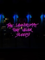 Neon pink text reads 'The Laundromat That Never Sleeps' against a dark background. The illumination casts a vibrant glow, suggesting a bustling, urban nightlife setting. The surrounding area appears dimly lit, with hints of reflective surfaces.