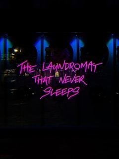 Neon pink text reads 'The Laundromat That Never Sleeps' against a dark background. The illumination casts a vibrant glow, suggesting a bustling, urban nightlife setting. The surrounding area appears dimly lit, with hints of reflective surfaces.