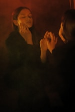 A person stands in front of a mirror with eyes closed, hands gently touching their face, creating a reflective and introspective scene. The lighting is dim and warm, casting soft shadows.
