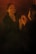 A person stands in front of a mirror with eyes closed, hands gently touching their face, creating a reflective and introspective scene. The lighting is dim and warm, casting soft shadows.