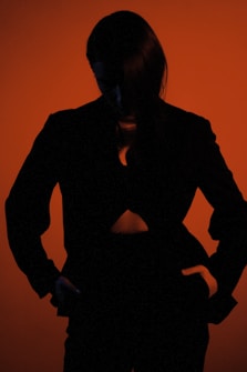 A silhouette of a person stands against a vivid orange background, partially obscured by shadow, creating a dramatic and mysterious atmosphere.