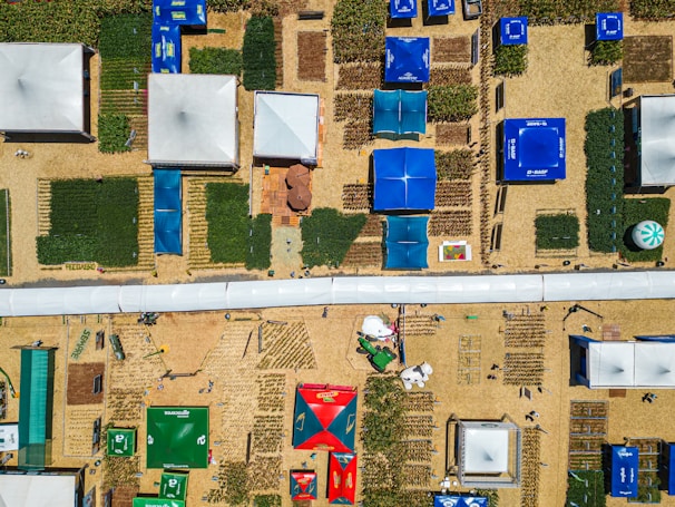 Aerial view of an organized outdoor area featuring various structures such as tents and exhibition pavilions. Rectangular plots of crops or greenery are interspersed between the structures, and colorful tarps providing shade are prominently visible. The pathways are filled with people walking around, and several logos and branding elements are present on the tents.