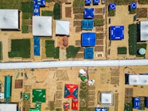 Aerial view of an organized outdoor area featuring various structures such as tents and exhibition pavilions. Rectangular plots of crops or greenery are interspersed between the structures, and colorful tarps providing shade are prominently visible. The pathways are filled with people walking around, and several logos and branding elements are present on the tents.