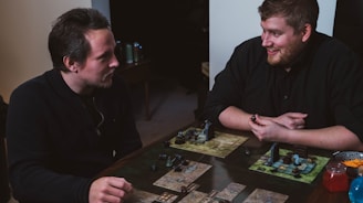 Two people are engaged in a tabletop game, sitting at a table with board game elements such as maps and figurines. The setting is casual and both individuals appear focused and enjoying the game.