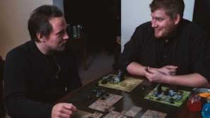 Two people are engaged in a tabletop game, sitting at a table with board game elements such as maps and figurines. The setting is casual and both individuals appear focused and enjoying the game.