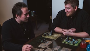 Two people are engaged in a tabletop game, sitting at a table with board game elements such as maps and figurines. The setting is casual and both individuals appear focused and enjoying the game.