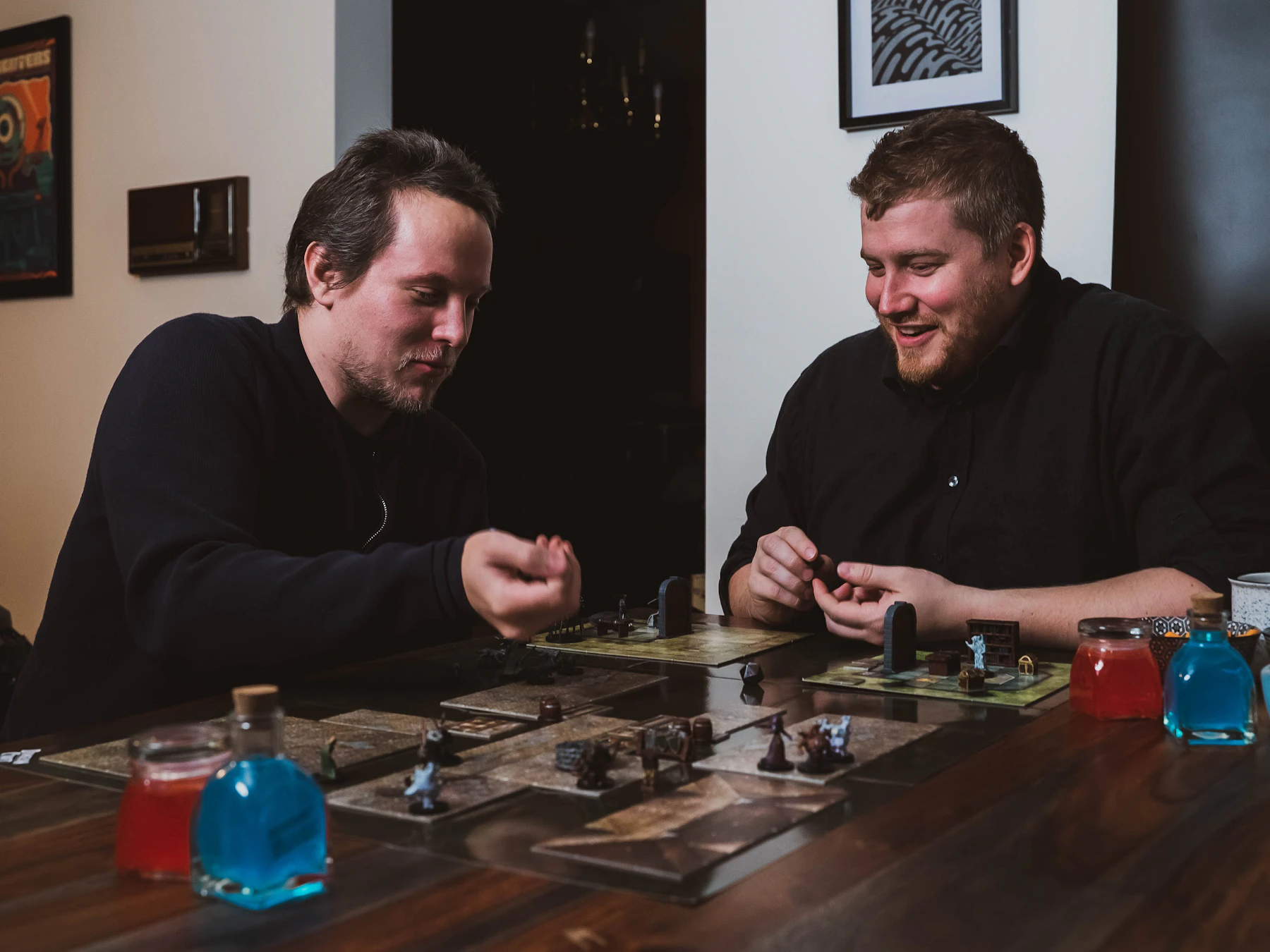 Two players sitting at a table in the middle of a tabletop game, with miniatures, map tiles and potion-bottle props laid out between them