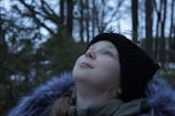 A young person gazes upwards in a serene outdoor forest setting. They are wearing a black knit hat adorned with a small fern and a coat with a fuzzy collar.