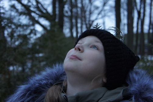 A young person gazes upwards in a serene outdoor forest setting. They are wearing a black knit hat adorned with a small fern and a coat with a fuzzy collar.