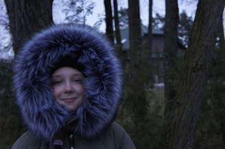 A person is wearing a large, fluffy hooded jacket, surrounded by trees. The hood is lined with thick gray fur, and the background features a forest setting with a house partially visible in the distance. The scene appears cold and wintry.