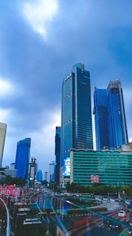 A bustling cityscape with towering skyscrapers including a prominent building displaying the BCA logo. The street below is filled with traffic and there are modern structures with glass windows. The overcast sky adds a moody ambiance to the scene.