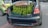 A car with its trunk open is parked on the street. Fresh green petai pods are hanging from the trunk, displayed for sale. A sign inside the trunk mentions pricing details in Malaysian Ringgit. Nearby, another car is parked with a light green color.