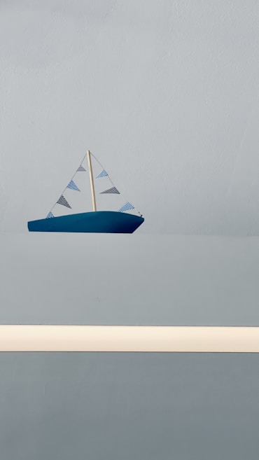 A small blue toy sailboat with a simple design and striped sails is positioned against a solid light gray background. The sails are triangular and feature alternating blue and black stripes. The overall scene is minimalistic, conveying a sense of calm and simplicity.