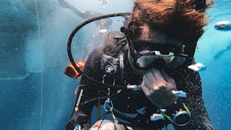 A scuba diver is underwater, wearing diving gear including a mask, wetsuit, and breathing apparatus. Bubbles surround the diver, who appears focused and engaged in adjusting equipment.