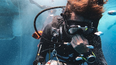 A scuba diver is underwater, wearing diving gear including a mask, wetsuit, and breathing apparatus. Bubbles surround the diver, who appears focused and engaged in adjusting equipment.