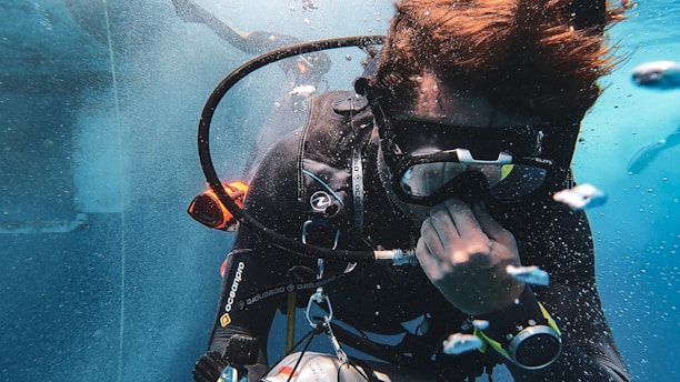 A scuba diver is underwater, wearing diving gear including a mask, wetsuit, and breathing apparatus. Bubbles surround the diver, who appears focused and engaged in adjusting equipment.