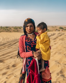 A woman holds a young child wrapped in a yellow blanket in a desert landscape. The woman is dressed in traditional, colorful clothing, accessorized with jewelry and a headscarf. Sand dunes stretch into the background under a clear blue sky, indicating a warm and arid environment.