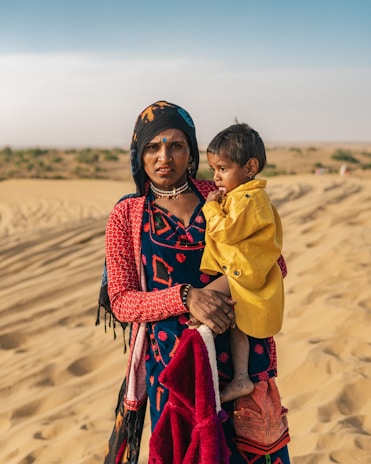 A Wayuu mother holding her child, both dressed in vibrant traditional clothing, with desert landscape in the background.