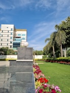 A modern building with a reflective glass facade stands adjacent to a lush, well-maintained green lawn bordered by a colorful array of flowers. Tall palm trees and other greenery line the garden. The sky is clear with a few clouds, and the scene is bathed in bright sunlight.