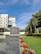 A modern building with a reflective glass facade stands adjacent to a lush, well-maintained green lawn bordered by a colorful array of flowers. Tall palm trees and other greenery line the garden. The sky is clear with a few clouds, and the scene is bathed in bright sunlight.