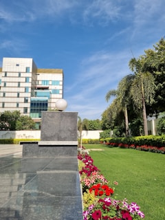 A modern building with a reflective glass facade stands adjacent to a lush, well-maintained green lawn bordered by a colorful array of flowers. Tall palm trees and other greenery line the garden. The sky is clear with a few clouds, and the scene is bathed in bright sunlight.
