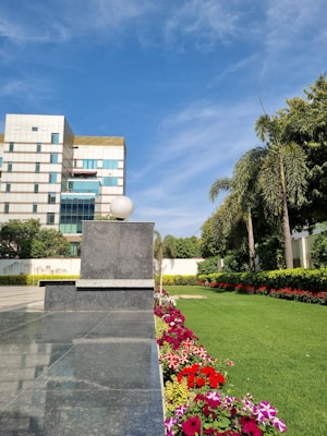A modern building with a reflective glass facade stands adjacent to a lush, well-maintained green lawn bordered by a colorful array of flowers. Tall palm trees and other greenery line the garden. The sky is clear with a few clouds, and the scene is bathed in bright sunlight.