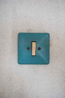 A light switch with a teal-colored plate mounted on a textured white wall.