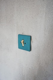 A single beige light switch mounted on a textured, light gray wall with a teal switch plate. The simplicity and minimalism of the setting emphasize a rustic or vintage aesthetic.