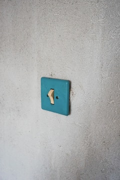 A single beige light switch mounted on a textured, light gray wall with a teal switch plate. The simplicity and minimalism of the setting emphasize a rustic or vintage aesthetic.