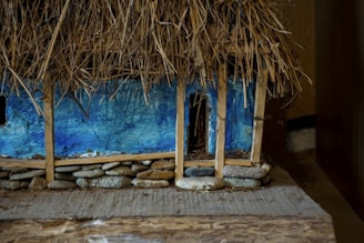 A rustic miniature model of a building constructed with a thatched roof made of straw, supported by wooden beams. The walls are painted in a vivid blue hue, and the base is lined with small, rounded stones. The detailed texture of straw and stones gives a natural and earthy feel to the model.
