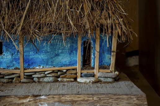 A rustic miniature model of a building constructed with a thatched roof made of straw, supported by wooden beams. The walls are painted in a vivid blue hue, and the base is lined with small, rounded stones. The detailed texture of straw and stones gives a natural and earthy feel to the model.