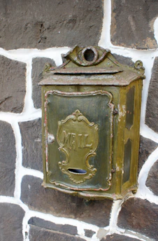 An antique metal mailbox with the word 'MAIL' embossed on the front, mounted on a stone wall. The mailbox has a rustic, weathered appearance, featuring a decorative design and an aged, greenish patina.