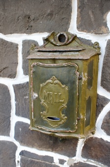 An antique metal mailbox with the word 'MAIL' embossed on the front, mounted on a stone wall. The mailbox has a rustic, weathered appearance, featuring a decorative design and an aged, greenish patina.