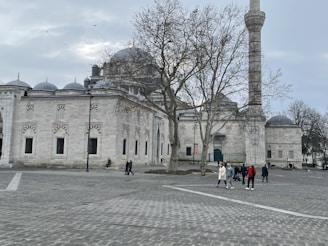 A historic mosque with an imposing structure made of light-colored stone features a central dome flanked by smaller domes and a tall minaret. The architecture includes intricate designs on the windows and walls. In the foreground, a large open plaza with cobblestone paving is visible, where a few people walk leisurely. Bare trees and a cloudy sky suggest a chilly day.