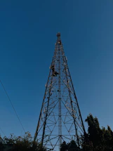 A tall metal telecommunications tower set against a clear blue sky. The structure is lattice-like with crisscrossing metal beams and extends high above the surrounding trees. The base is surrounded by foliage, and a few utility lines are visible.