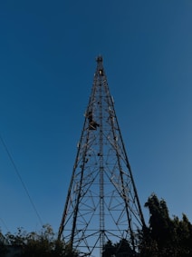 A tall metal telecommunications tower set against a clear blue sky. The structure is lattice-like with crisscrossing metal beams and extends high above the surrounding trees. The base is surrounded by foliage, and a few utility lines are visible.
