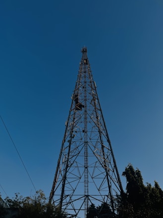 A tall metal telecommunications tower set against a clear blue sky. The structure is lattice-like with crisscrossing metal beams and extends high above the surrounding trees. The base is surrounded by foliage, and a few utility lines are visible.