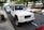 A white off-road SUV is parked on the street alongside several other cars. The vehicle has a rugged appearance with large tires and a robust front bumper. Trees and buildings can be seen in the background, adding a suburban feel to the scene.