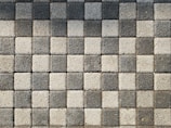 A pattern of square pavers arranged in a checkered layout with alternating light and dark shades of gray. The surface texture of each paver is rough and pebble-like.