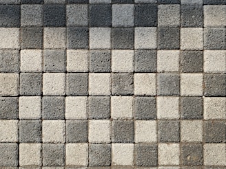 A pattern of square pavers arranged in a checkered layout with alternating light and dark shades of gray. The surface texture of each paver is rough and pebble-like.