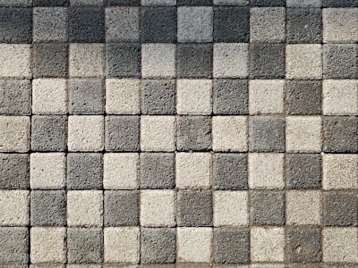 A pattern of square pavers arranged in a checkered layout with alternating light and dark shades of gray. The surface texture of each paver is rough and pebble-like.