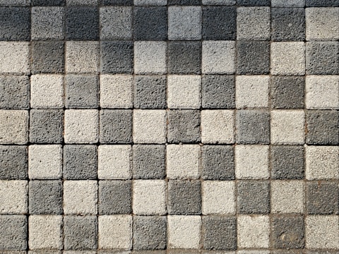 A pattern of square pavers arranged in a checkered layout with alternating light and dark shades of gray. The surface texture of each paver is rough and pebble-like.