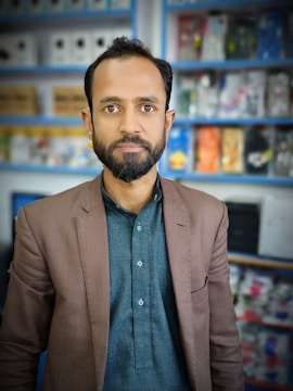 A man with a beard is standing indoors, wearing a brown blazer over a dark shirt. Behind him are shelves stocked with various items, possibly electronics or household goods, displayed in a retail setting. The background is slightly blurred, focusing attention on the man.